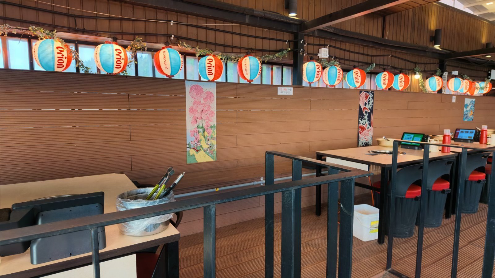 Restaurant interior in pale wood with a row of tables along a brick wall, paper lanterns strung across the ceiling and an ordering tablet at each place.