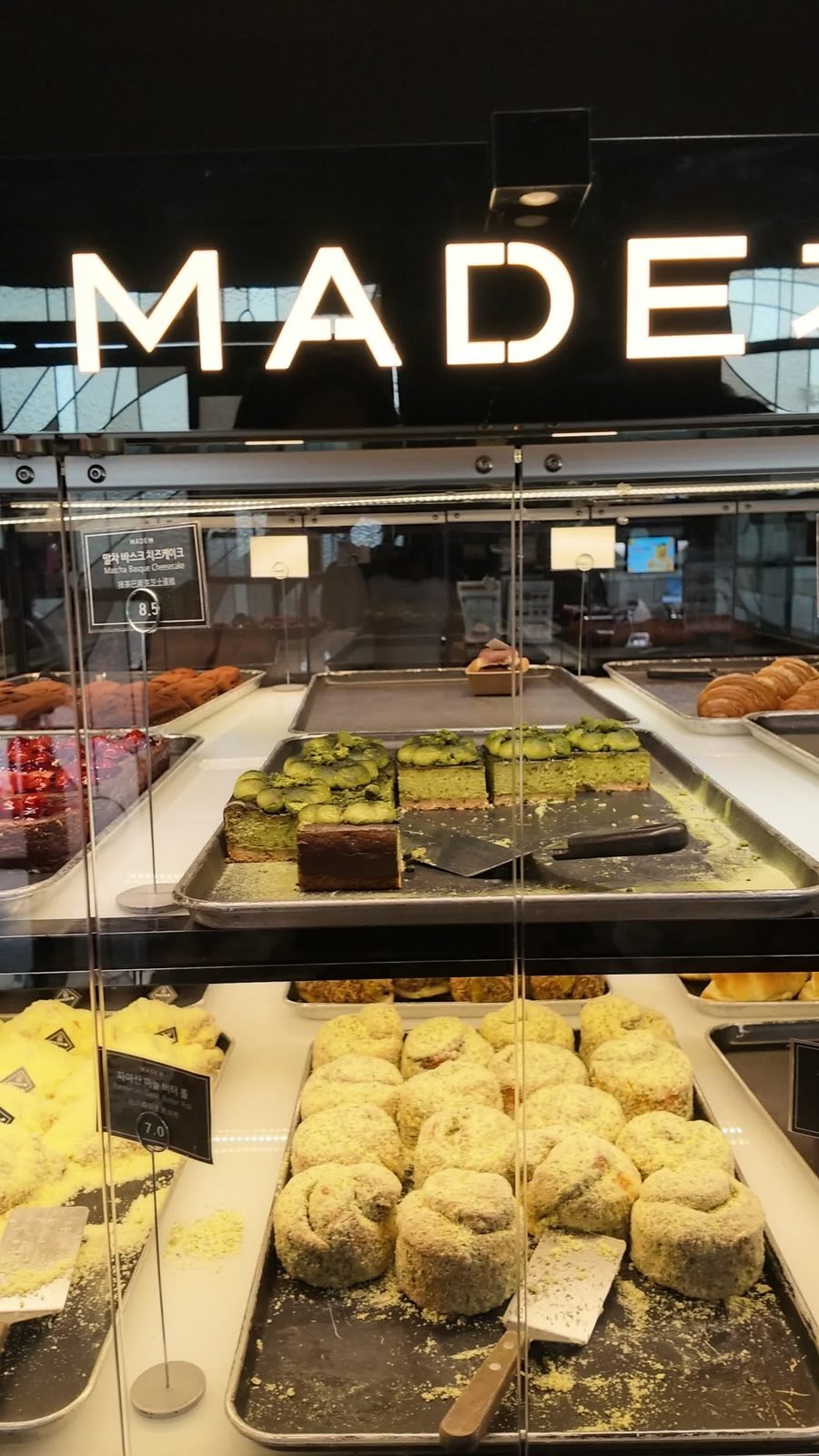 Bakery display case with sliced green matcha basque cheesecake on a tray, a price tag reading Matcha Basque Cheesecake 8.5, croissants and green-dusted rolls on the shelf below