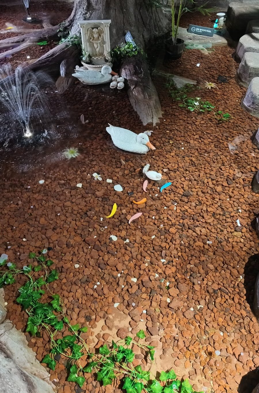 Indoor pond with red gravel, fountain jets, duck ornaments and a small white angel relief at the base of a tree trunk
