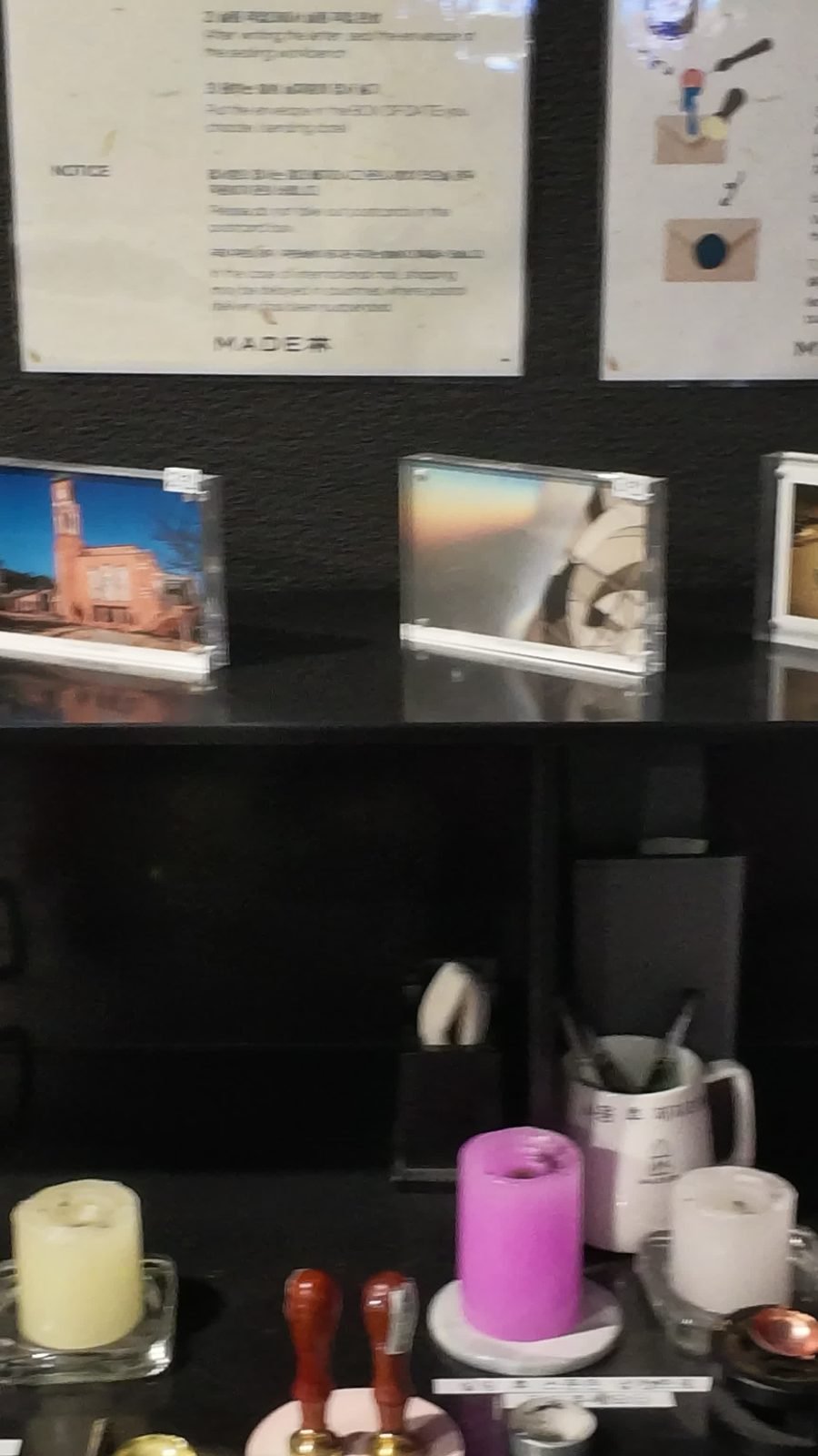 Wax seal station with two wooden-handled brass stamps, a pink candle, cream candles and a melting spoon on a black counter, with framed postcards and a bilingual notice above