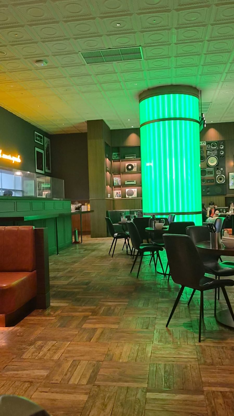 Interior of MJ23 with a tall backlit green column, wood flooring, dark green booth seating and empty tables.
