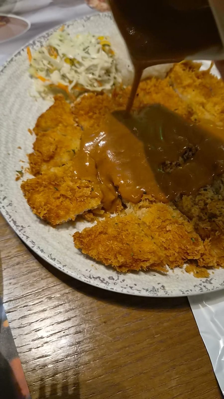 Sliced breaded pork cutlet on a wide speckled plate with brown sauce ladled over the middle, rice underneath and shredded cabbage coleslaw at the edge.
