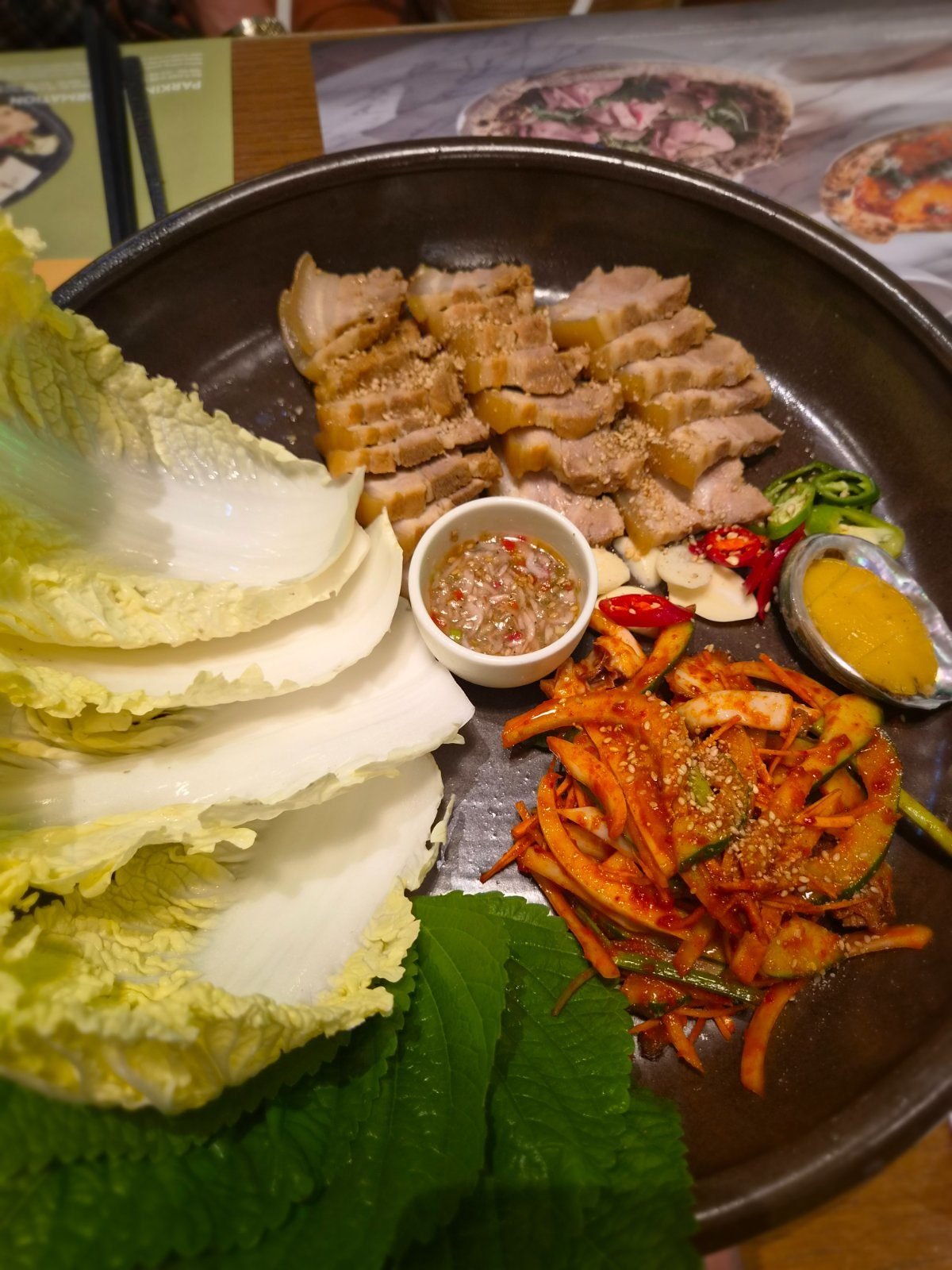A platter of thick-sliced boiled pork with napa cabbage leaves, perilla leaves, garlic, green chilli, a mustard dip and a mound of red dressed vegetables.