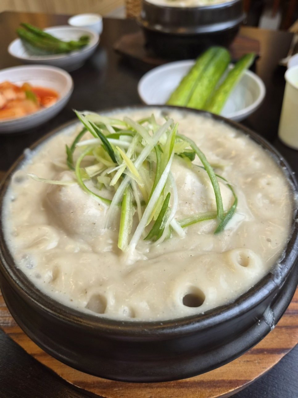 Deulkkae samgyetang in a black stone pot, thick opaque perilla broth topped with shredded scallion, with kimchi, whole cucumbers and green chillies alongside.