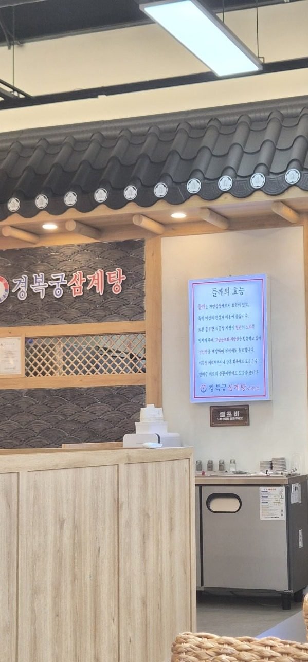 Tiled hanok roofline over the counter at Gyeongbokgung Samgyetang Yeongjong, with a wall notice about perilla seed and a wooden sign reading 셀프 바 above the self-service bar.