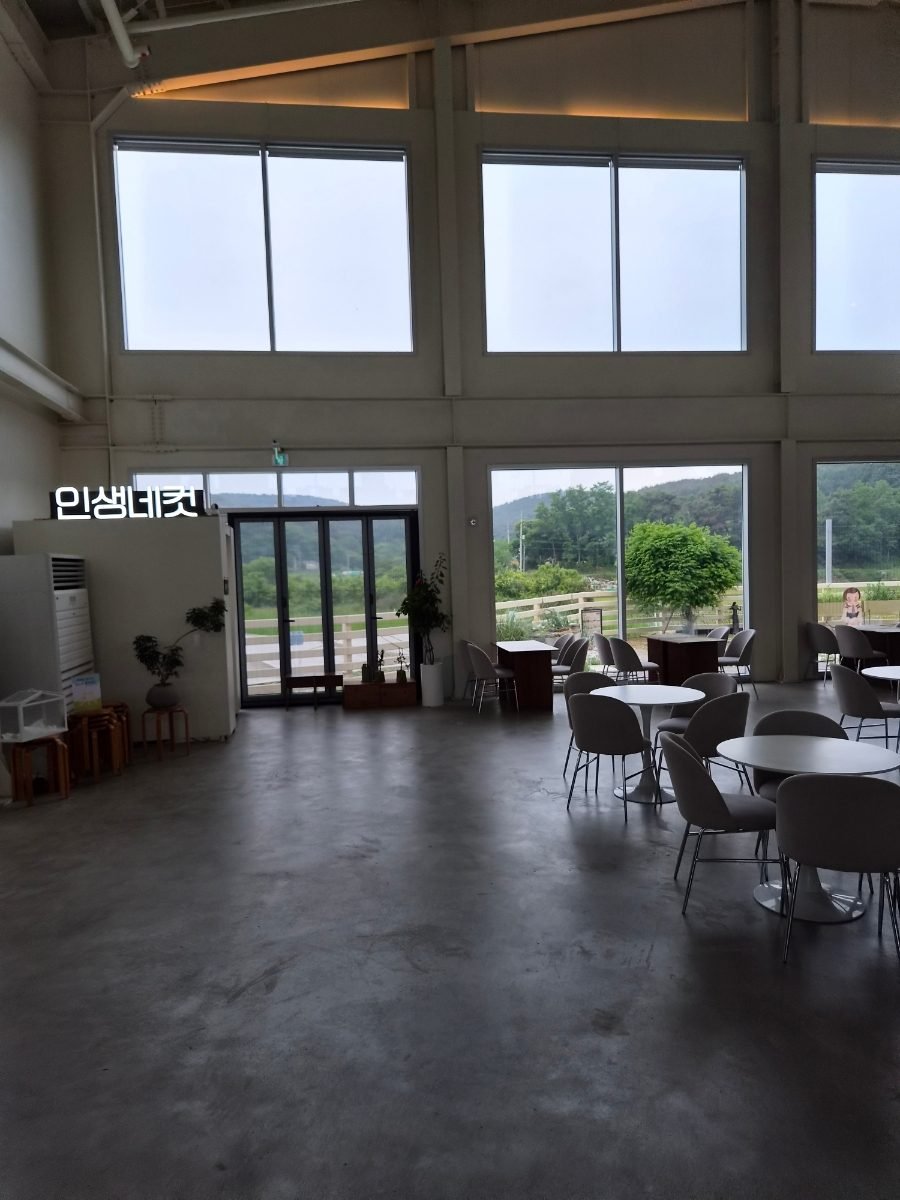 The cafe interior: a double-height room with tall windows, bare concrete floor, round white tables and grey chairs, and green fields visible outside.
