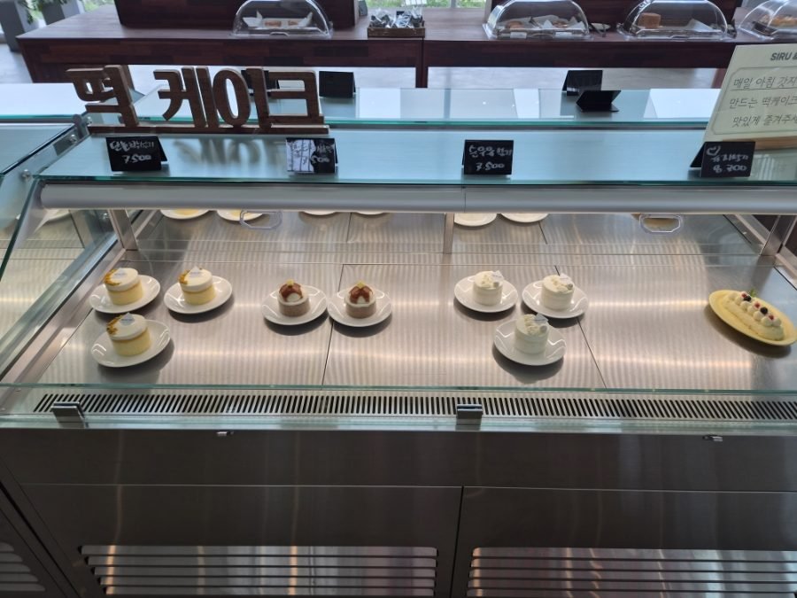 A glass showcase of individual rice cakes on white plates, with wooden Korean letters reading tteok cake standing on top and a price tag showing 7,500 won.