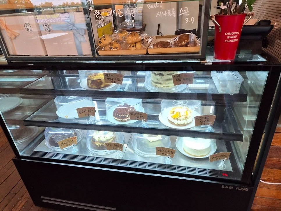 Glass chilled cake case holding cheesecakes, tarts and a brownie with handwritten Korean price labels, and bagged scones on the shelf above