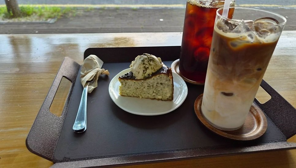 Slice of matcha basque cheesecake topped with cream on a white plate, beside an iced americano and an iced latte on a dark tray at a window table