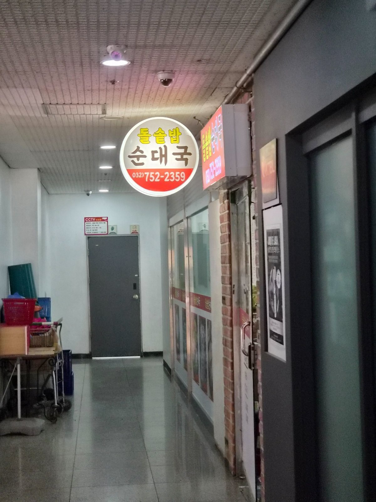 Illuminated round sign reading 돌솥밥 순대국 hanging in an interior building corridor with a dark entrance door below it