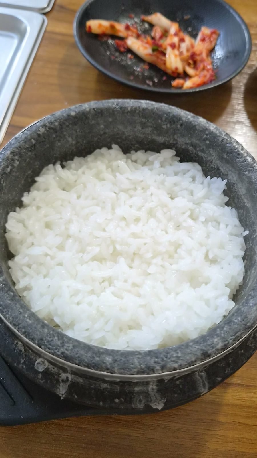 White rice cooked in a heavy grey stone pot on a wooden table, with a plate of fresh cabbage kimchi behind it