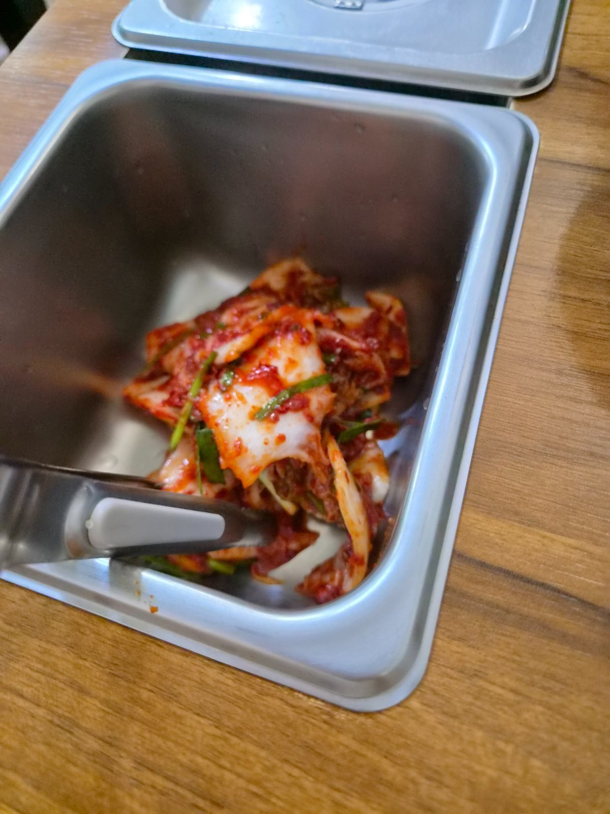 Cabbage kimchi in a stainless serving tub on a wooden counter with tongs resting inside