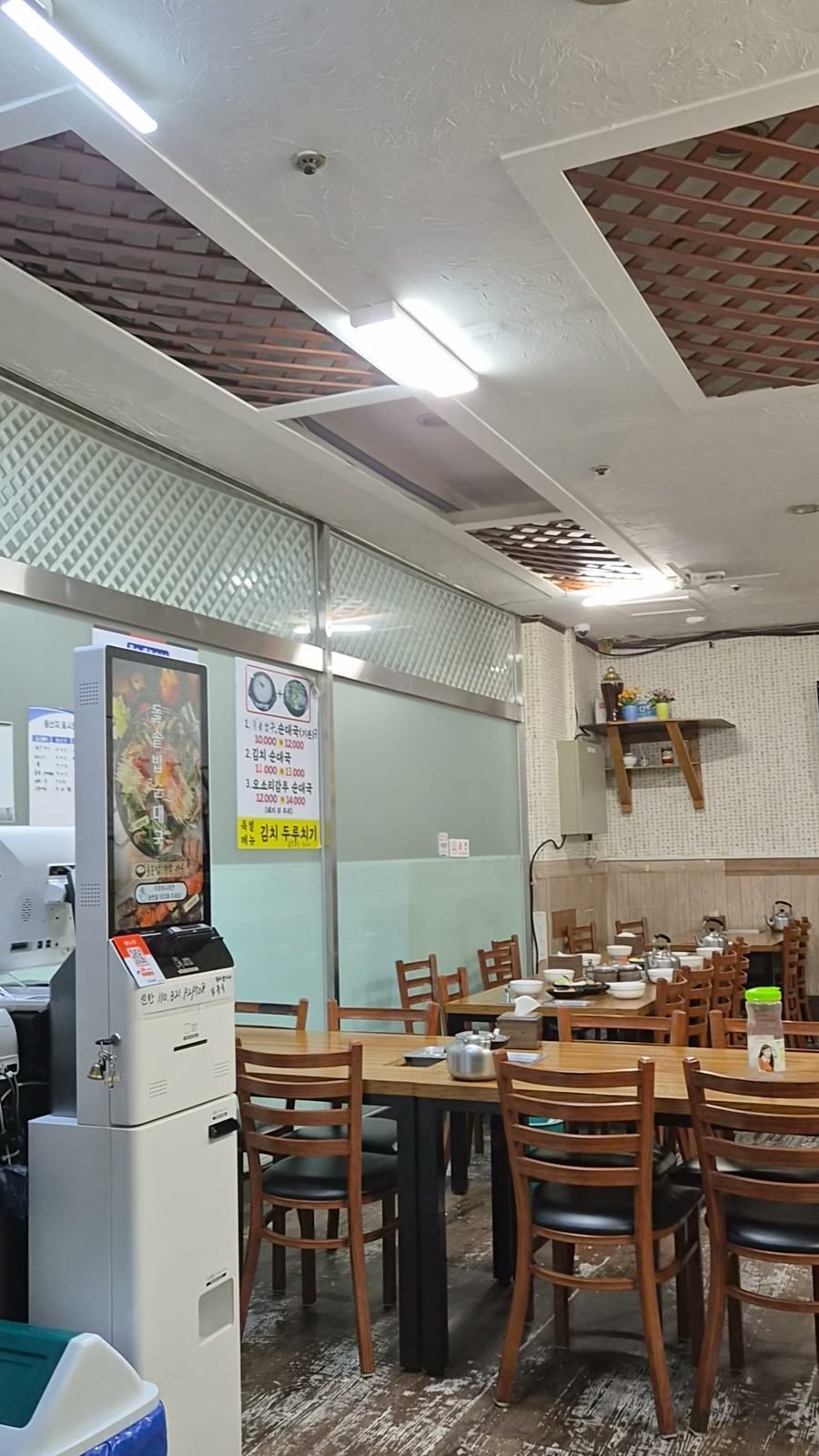 Interior dining room with plain wooden tables and chairs, metal kettles and stainless bowls set out, and a menu sheet taped to the partition wall