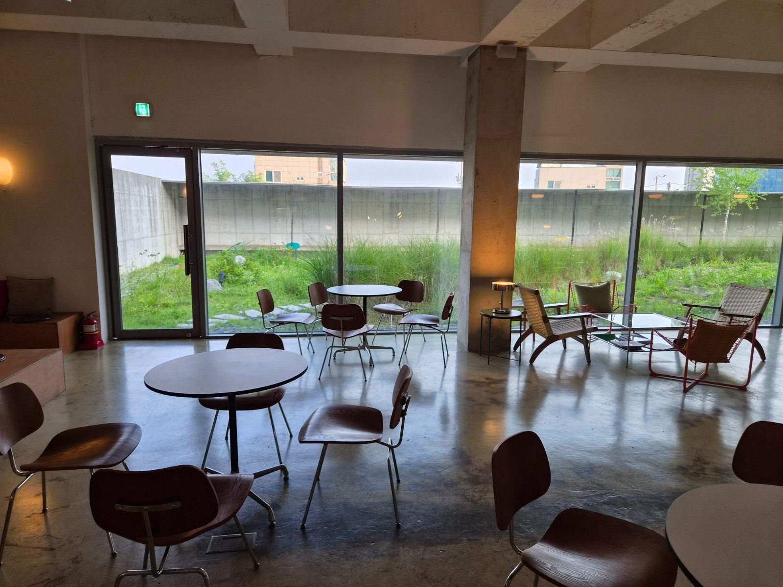 High ceilinged cafe interior with concrete columns, wood chairs and a glass wall onto a grass garden