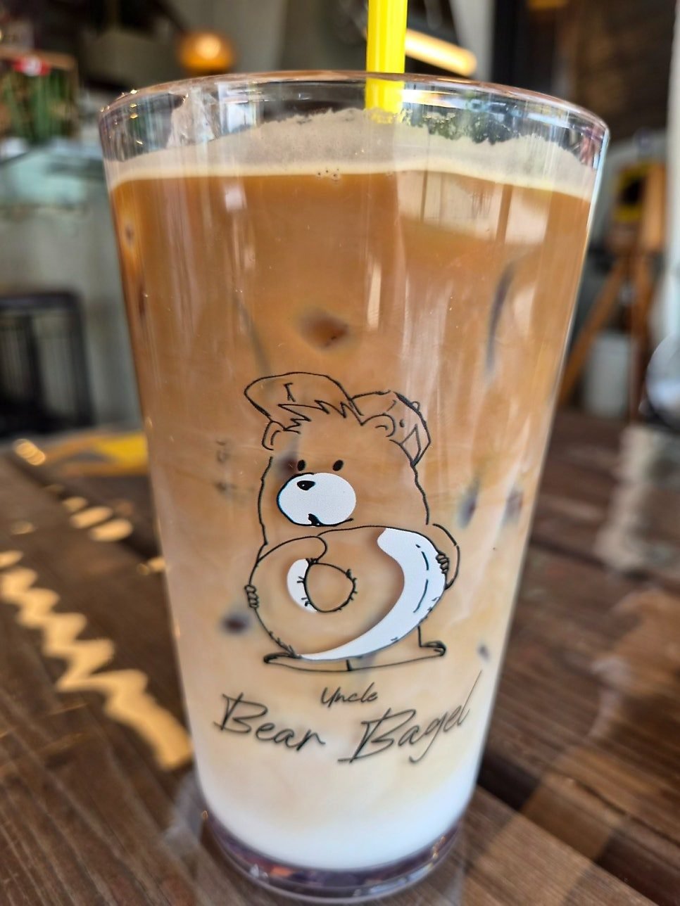 Iced latte in a tall glass printed with a cartoon bear and the words Uncle Bear Bagel, milk and coffee in separate layers