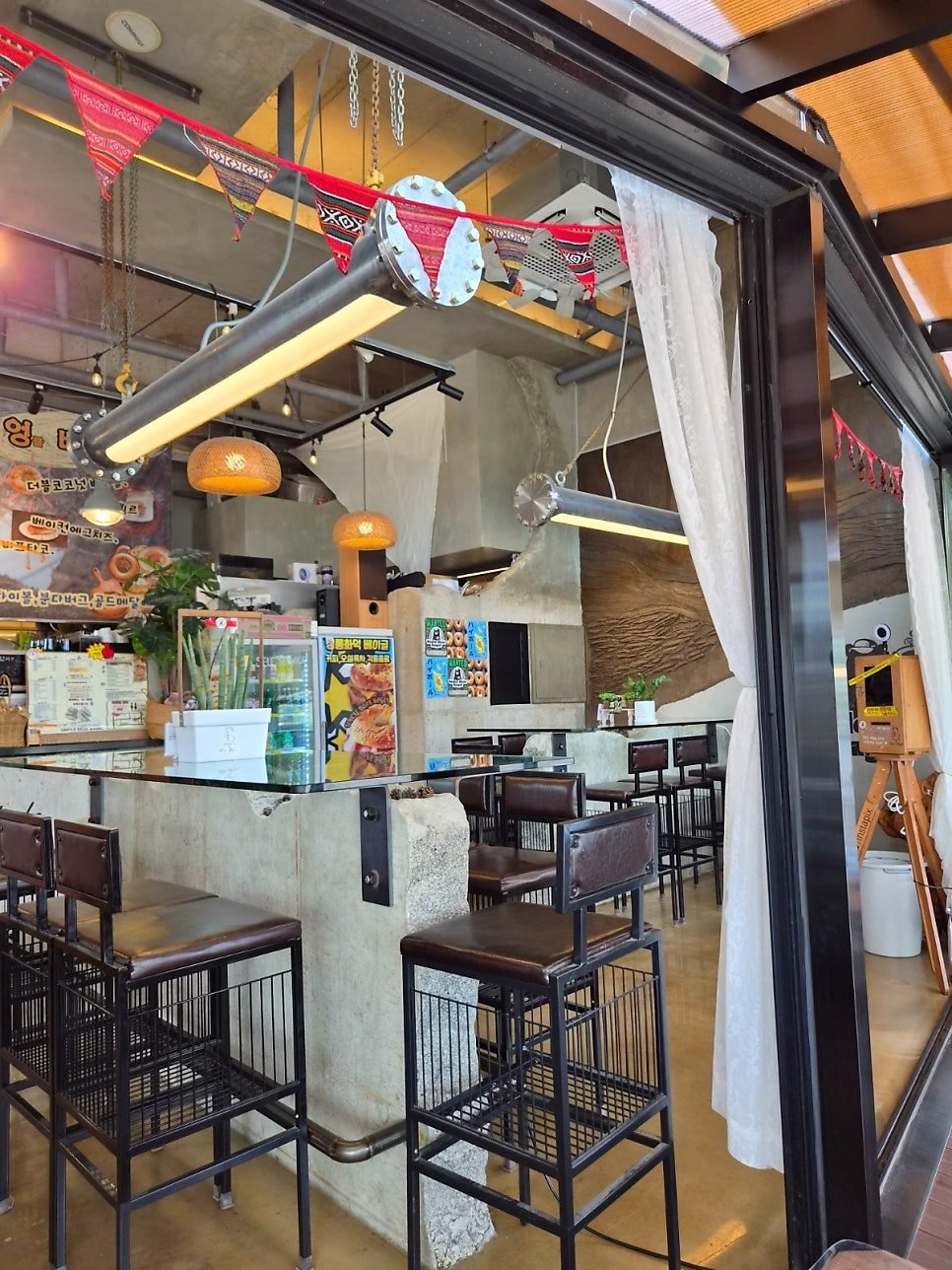 Exposed concrete interior with high chairs at a bar counter, pendant lights and a bunting garland, seen through the window