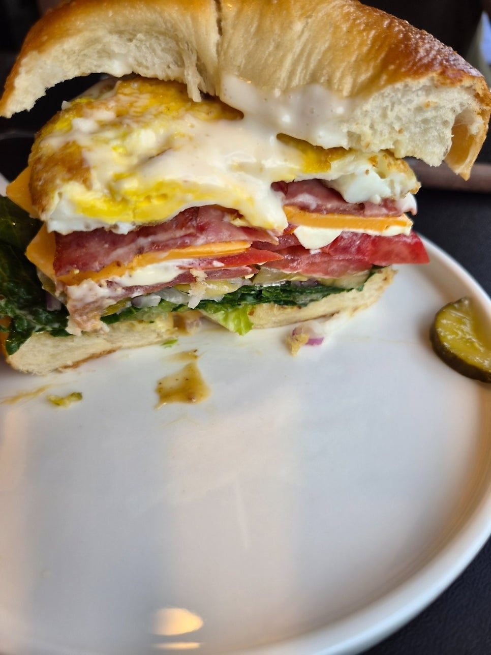 Cross section of a bagel sandwich with a soft egg yolk running over bacon, cheese, tomato and lettuce, with a pickle slice on the plate