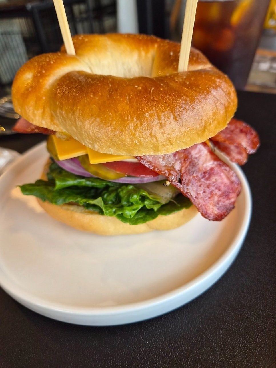 Bagel sandwich held together with a skewer, stacked with sausage, bacon, cheddar, lettuce, red onion and tomato on a white plate