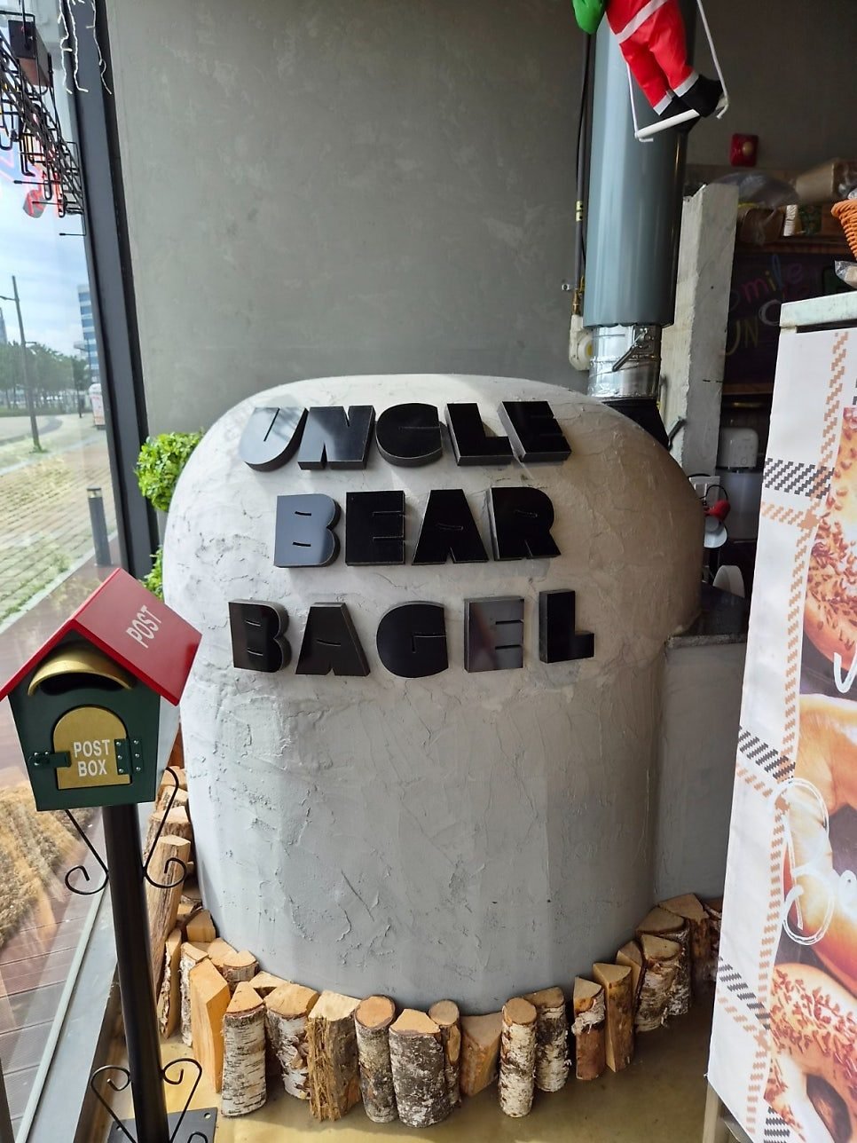 Large stone bagel sculpture spelling UNCLE BEAR BAGEL beside a red postbox and stacked logs at the shop entrance